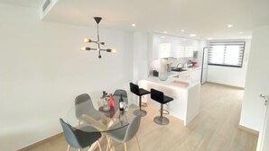Una cocina moderna con armarios blancos y una isla central, un comedor con una mesa de vidrio y sillas negras, y un colgante de techo.