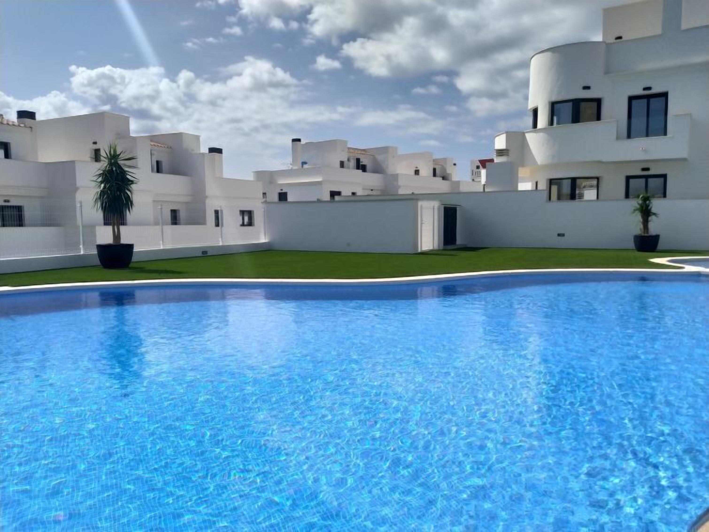 A modern pool area with a clear blue pool, surrounded by white buildings with balconies and potted plants.