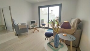 Una sala moderna con un sofá, una silla, una mesita y una lámpara. Hay un ordenador portátil sobre un soporte y un jarrón con flores secas en la mesa.