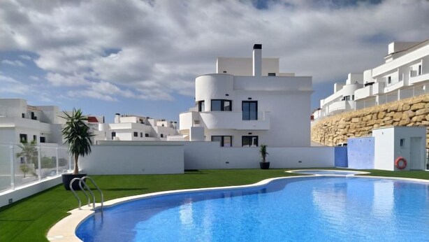 Un área de piscina moderna con una piscina azul cristalina, rodeada de edificios blancos y un muro de piedra.