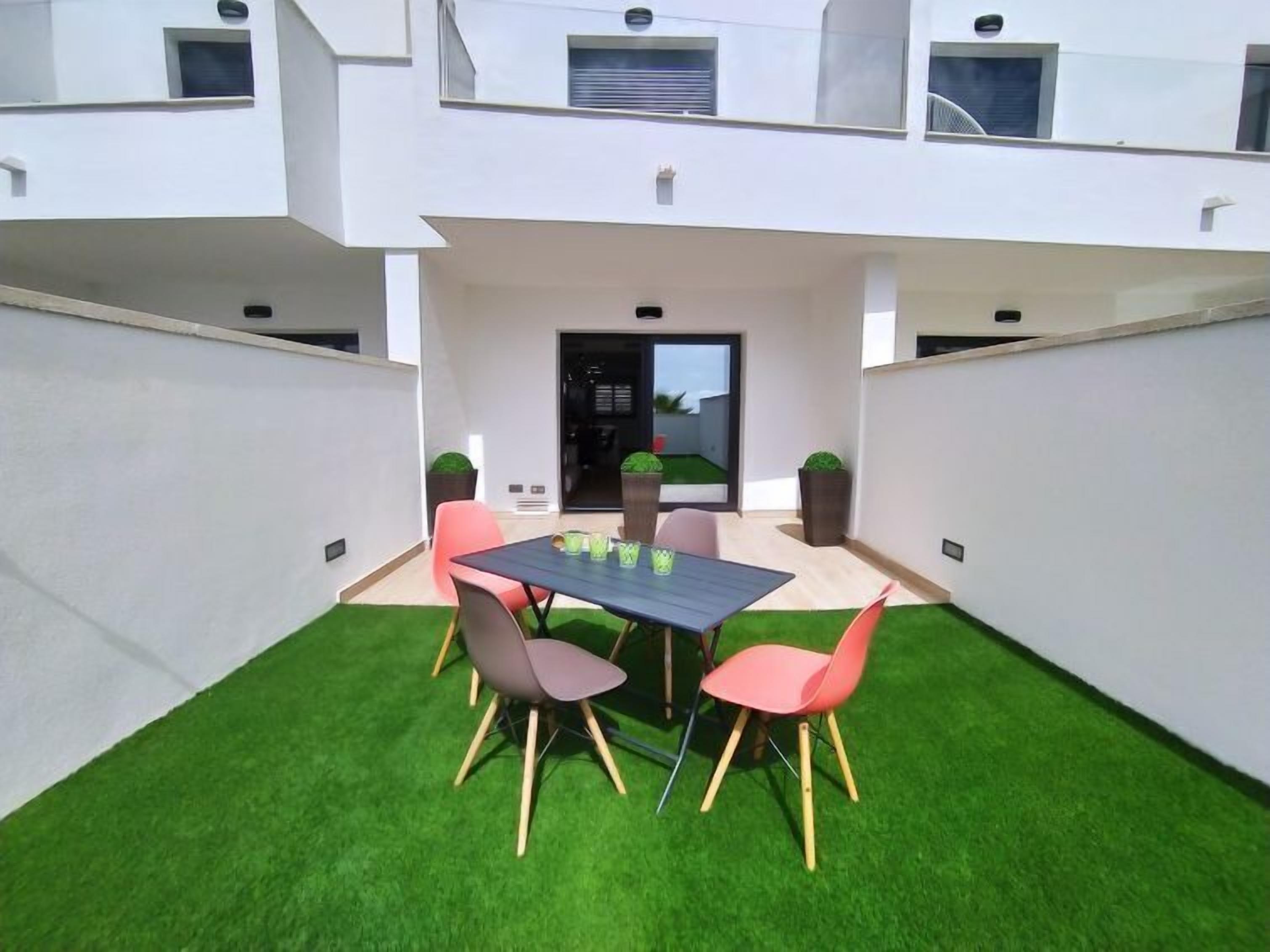 A modern outdoor patio with a dining table and chairs, surrounded by artificial grass and potted plants.