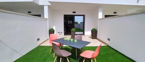 A modern outdoor patio with a dining table and chairs, surrounded by artificial grass and potted plants.