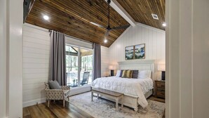 A bedroom with a wooden ceiling, a large bed with white bedding, a bench, a chair, and a window with curtains. - Platinum Ridge - Luxury One Bedroom with Bunk Nook (Broken Bow)