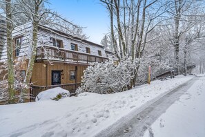 Um edifício coberto de neve com varanda, rodeado por árvores e um caminho nevado.