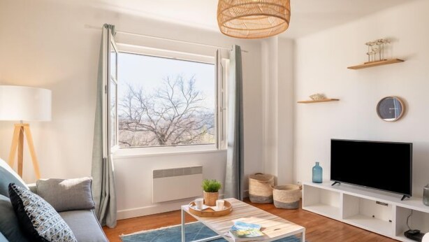 Ein modernes Wohnzimmer mit einer Couch, einem Couchtisch, einem Fernseher und einem großen Fenster mit Vorhängen.