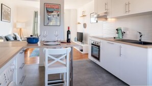 Eine moderne Küche mit weißen Schränken, einem eingebauten Backofen und einer Kaffeemaschine. Es gibt eine Essecke mit einem Tisch, der für zwei Personen gedeckt ist, und einem weißen Stuhl. Auf dem Tisch stehen eine Weinflasche und Gläser.