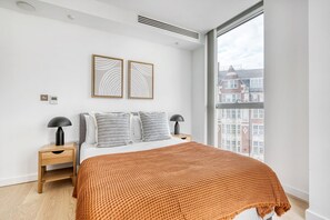 A modern bedroom with a large window, a bed with a textured orange blanket, two bedside tables with lamps, and two framed wall art pieces.