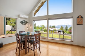 Un coin repas avec une table et des chaises, de grandes fenêtres offrant une vue sur la verdure et les montagnes, et une horloge fixée au mur.
