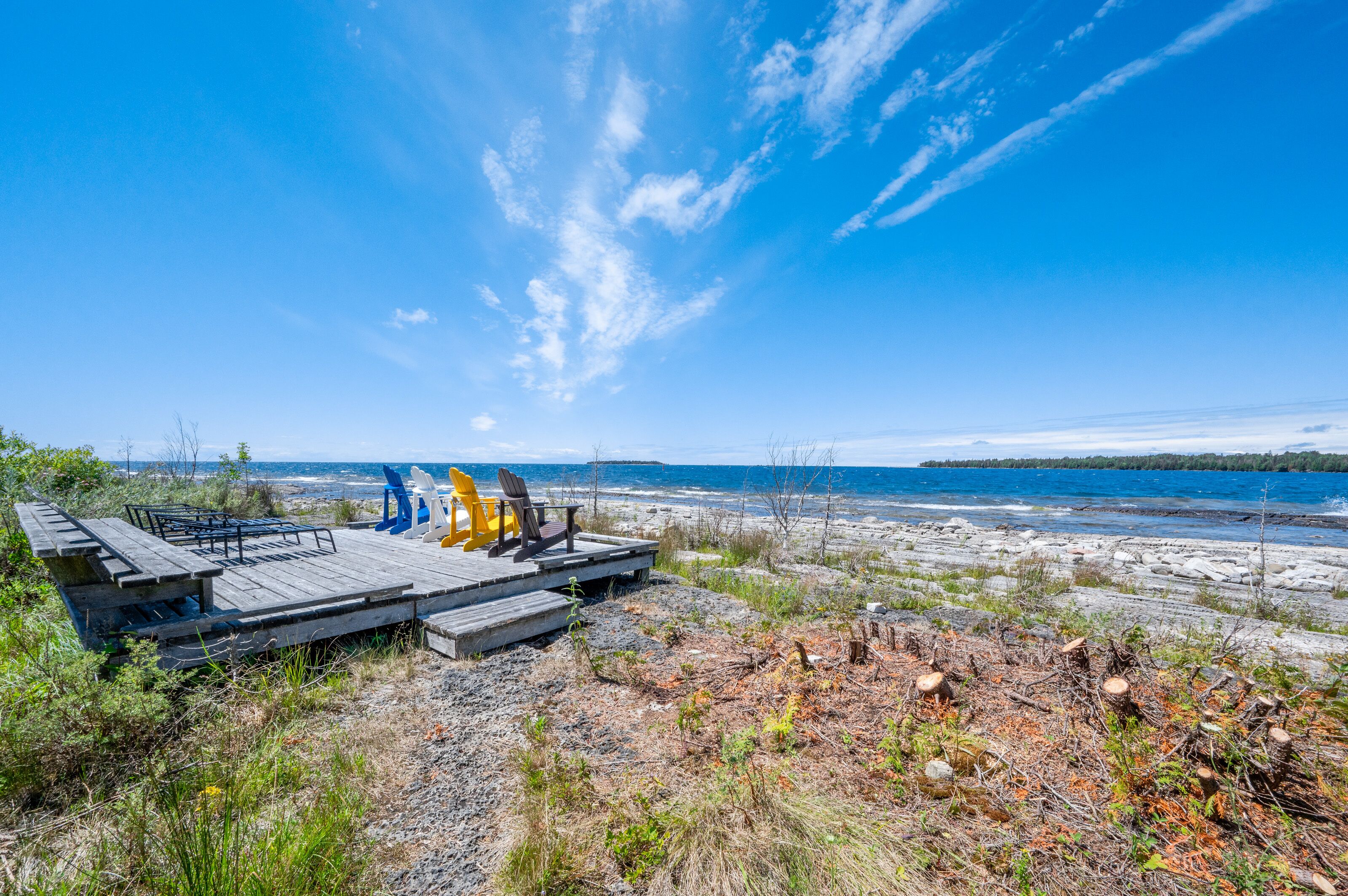 Een houten terras met Adirondack-stoelen, met uitzicht op een strand en de oceaan, met bomen in de verte.