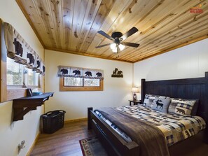Une chambre avec un plafond en bois, un lit recouvert d’une courtepointe à motifs, une table de chevet avec une lampe, une étagère fixée au mur et une fenêtre donnant sur un paysage.