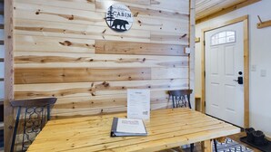 Interior de una cabaña con pared de madera, un cartel de bienvenida, una mesa, dos sillas y una puerta.