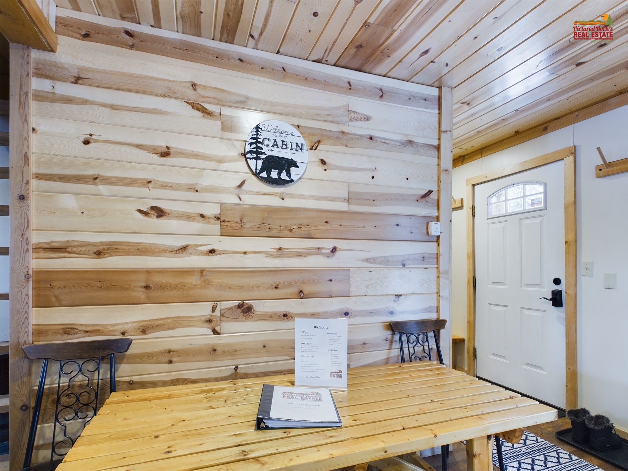 A cabin interior with a wooden wall, a welcome sign, a table, two chairs, and a door.