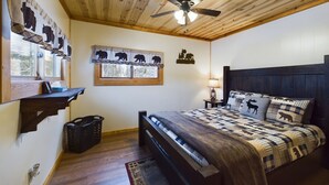 Un dormitorio con techo de madera, una cama con colcha estampada, una mesita de noche con lámpara, un estante fijado a la pared y una ventana con vistas.