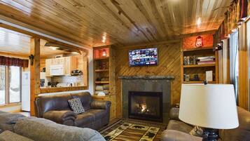 A cozy living room with a fireplace, leather sofas, a TV mounted on the wall, and wooden shelves with books and decorations.