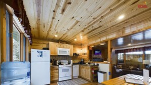 Una cocina acogedora de estilo cabaña, con armarios de madera, un microondas, una estufa y un lavabo. Cuenta con un dispensador de agua grande y una mesa de madera con bancos.