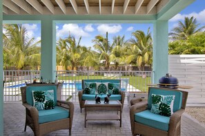 A covered patio area with wicker furniture, a pool, and palm trees in the background.