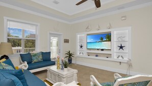 Ein Wohnzimmer mit einem blauen Sofa, weißen Sesseln, einem Couchtisch und einem Fernseher an der Wand.