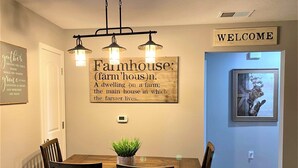 Een eetruimte met een houten tafel en stoelen, een bord met de tekst 'Welcome', een bord met de tekst 'Farmhouse' en een lijst met een schilderij van een koe.