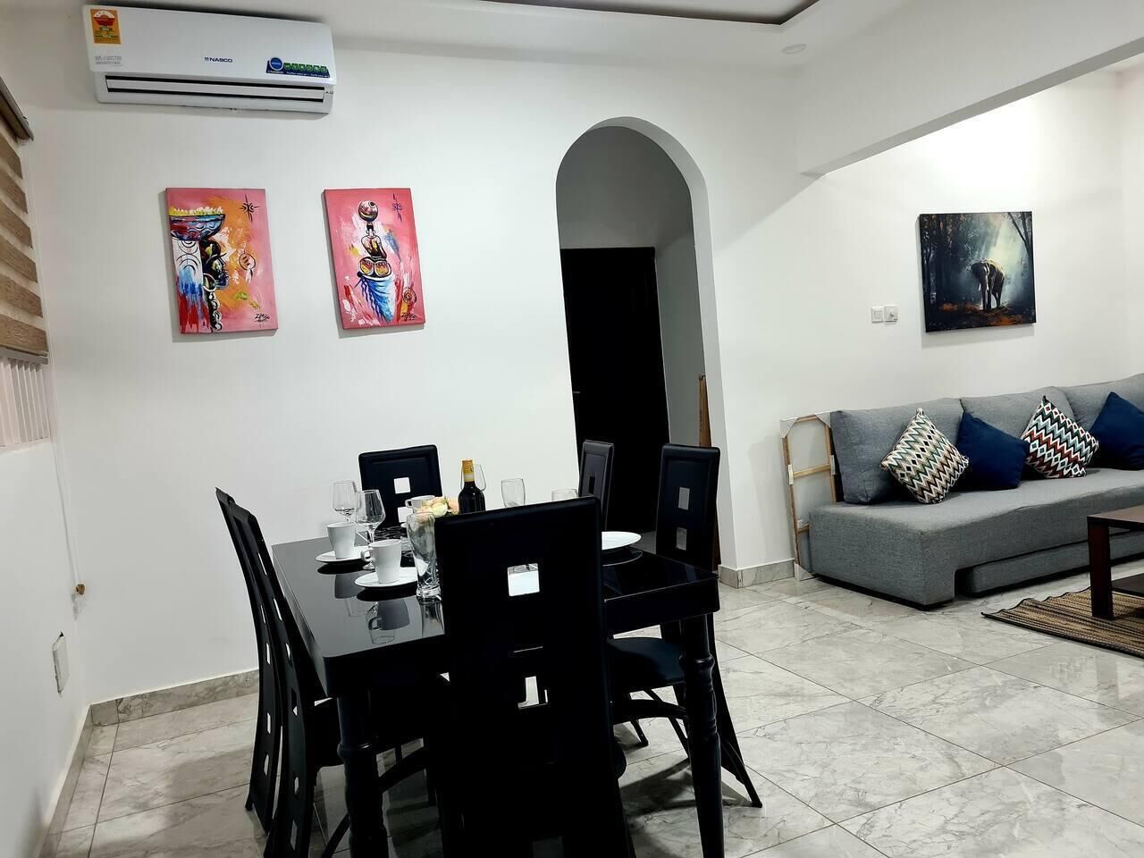 Un comedor con una mesa y sillas negras, un sofá gris con cojines estampados y dos cuadros en la pared.