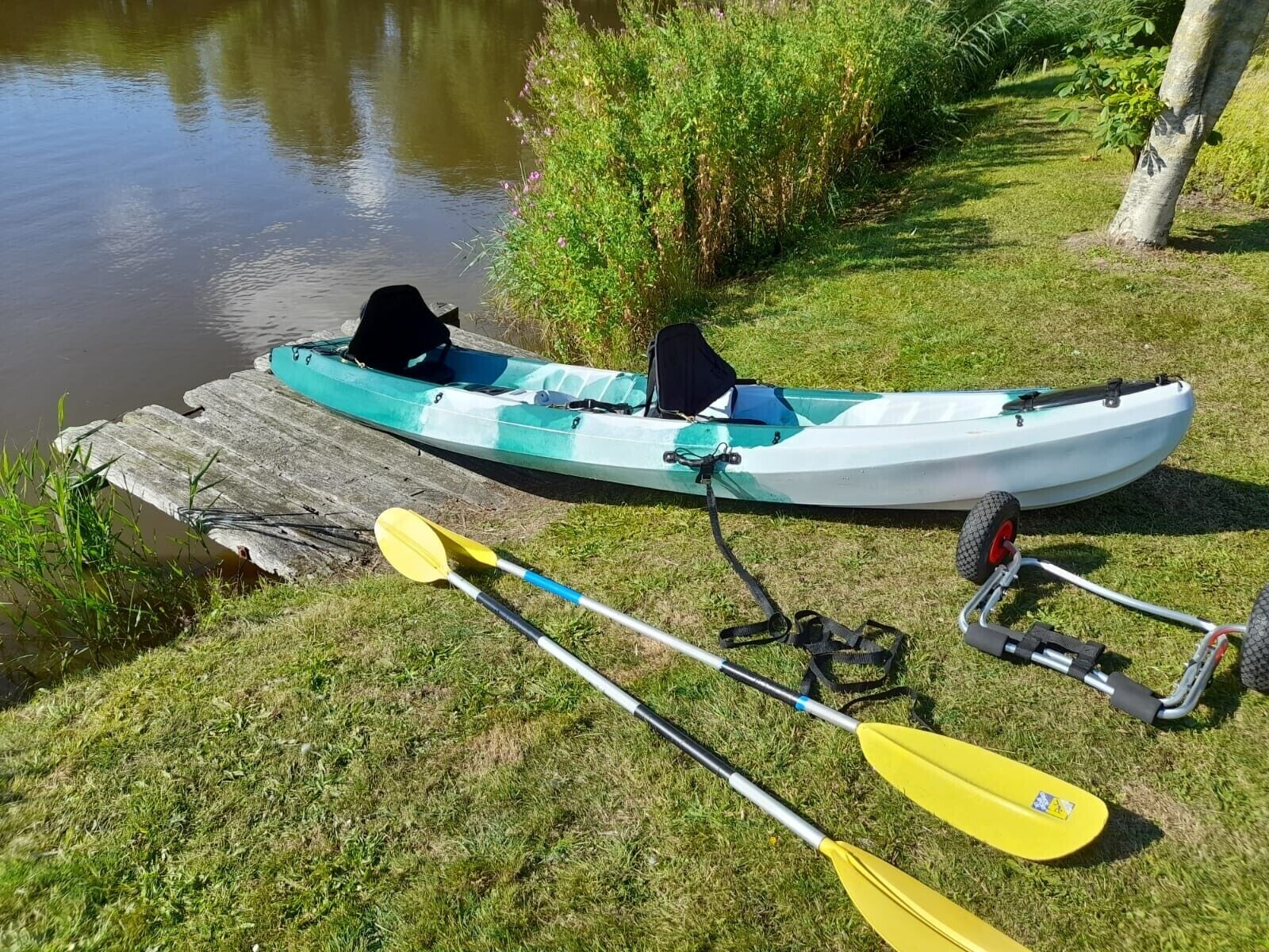 Een kajak afgemeerd aan een houten steiger, met twee peddels en een karabijnhaak in de buurt.