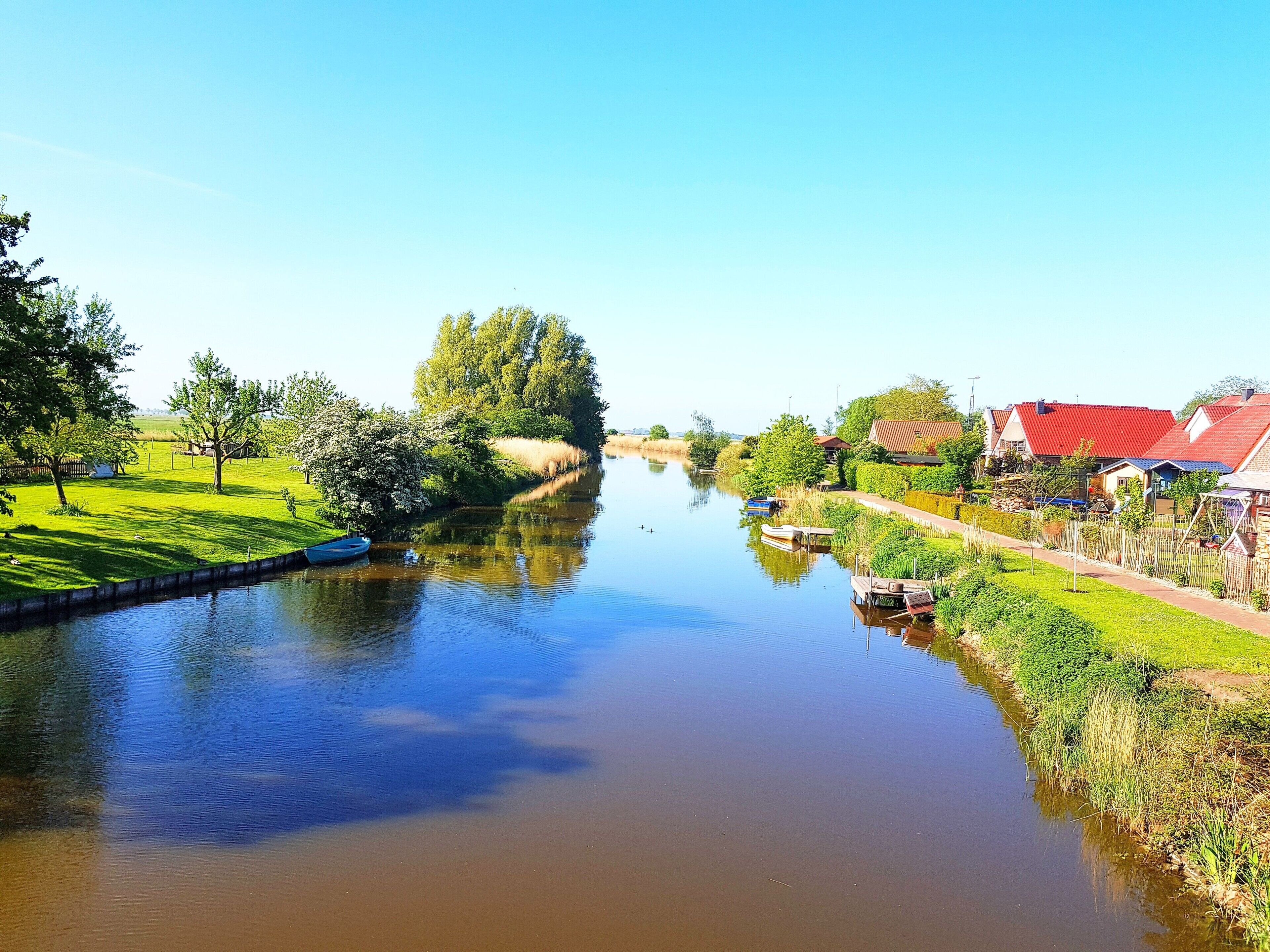 A calm river with houses on the right bank and a tree-lined path on the left.
