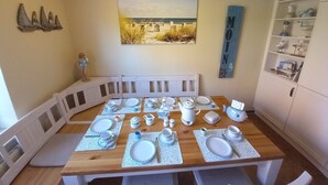 Een eetruimte met een houten tafel voor vier personen, omringd door witte stoelen. Een witte kast met glazen deuren staat aan de rechterkant.
