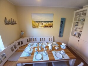 A dining area with a wooden table set for four, surrounded by white chairs. A white cabinet with glass doors is to the right.