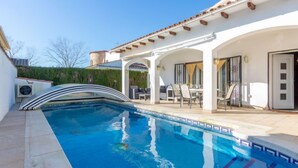 A swimming pool with a curved edge, a covered patio area with outdoor furniture, and a white building with a tiled roof. - Erbanozi by Interhome (Empuriabrava)