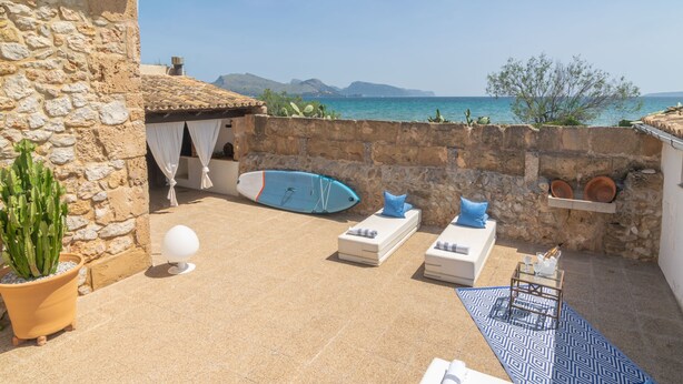 Une terrasse extérieure aux murs de pierre, avec des fauteuils de jardin blancs, une planche de surf bleue et un cactus en pot.