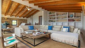 Un salon spacieux avec un plafond haut, un mur en pierre, un plafond en bois, un canapé blanc agrémenté de coussins bleus, une table basse et une bibliothèque.