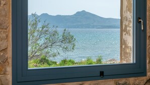 Une fenêtre donnant sur une mer calme, des montagnes lointaines et un arbre au premier plan.