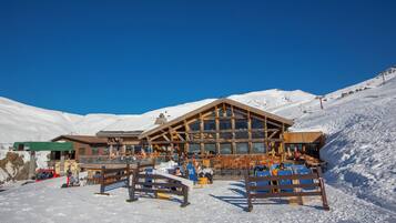 Un chalet en bois, avec un télésiège visible au loin, entouré de montagnes enneigées.