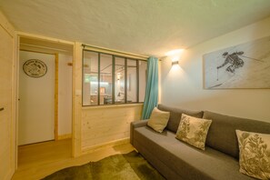 A cozy living room with a wooden floor, a grey sofa with palm leaf cushions, a window with a curtain, and a wall clock.