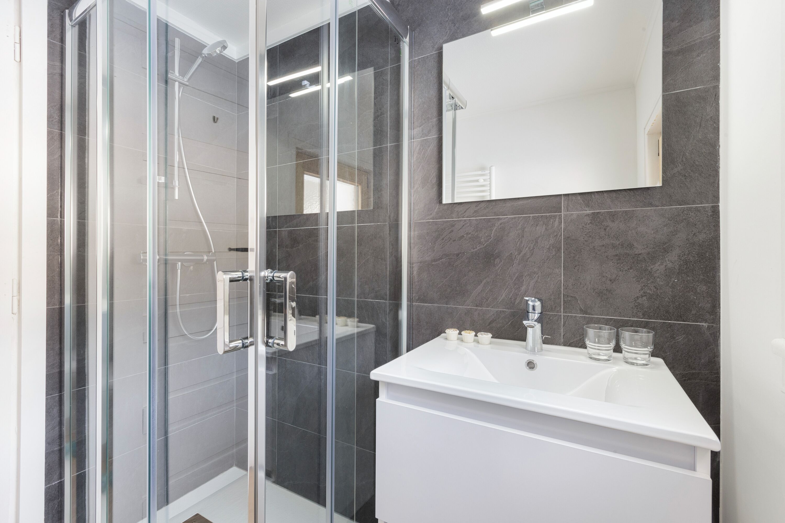 Een moderne badkamer met een douche in glazen cabine, een wit wastafelmeubel met twee glazen en een grote spiegel.