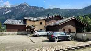 Un edifici amb zona d'aparcament, dos cotxes i un paisatge de muntanya al fons.