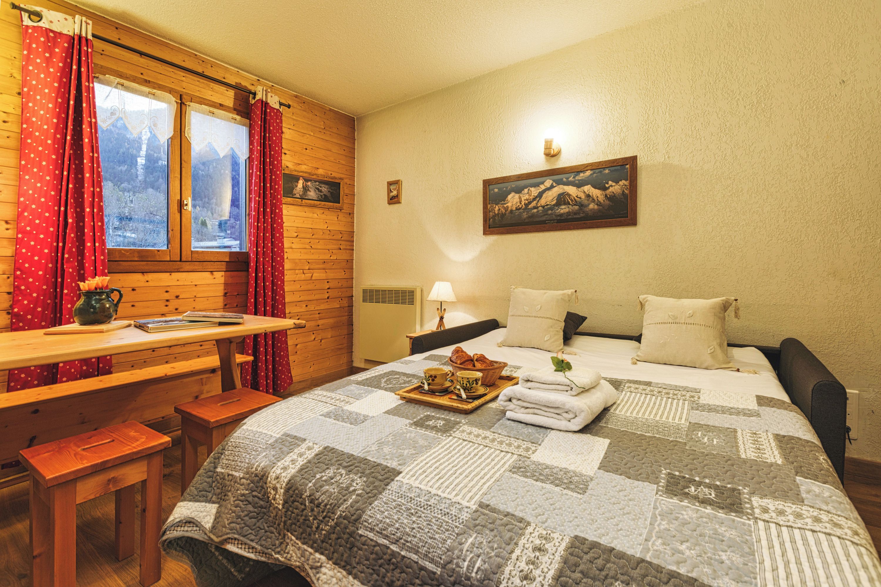 Een knus ingerichte kamer met een houten bureau, een bed met een sprei, een raam met gordijnen en een schilderij aan de muur.
