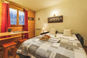 Una habitació acollidora amb escriptori de fusta, llit amb llençol de retalls, finestra amb cortines i un quadre al mur.