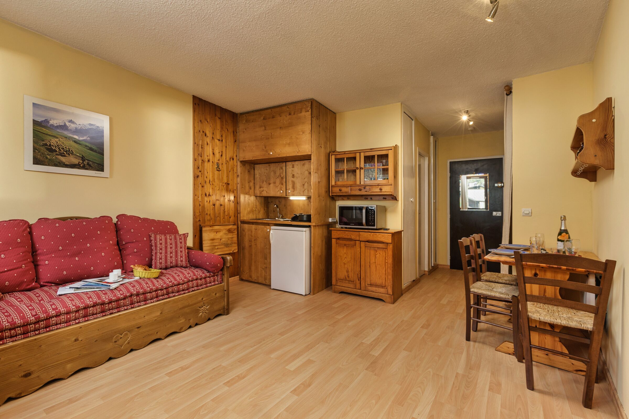 A cozy room with a wooden sofa, a dining table with chairs, a kitchen area with wooden cabinets, and a refrigerator.
