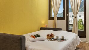 A hotel room with a bed, a breakfast tray, a window with curtains, and a view of a building and mountains.