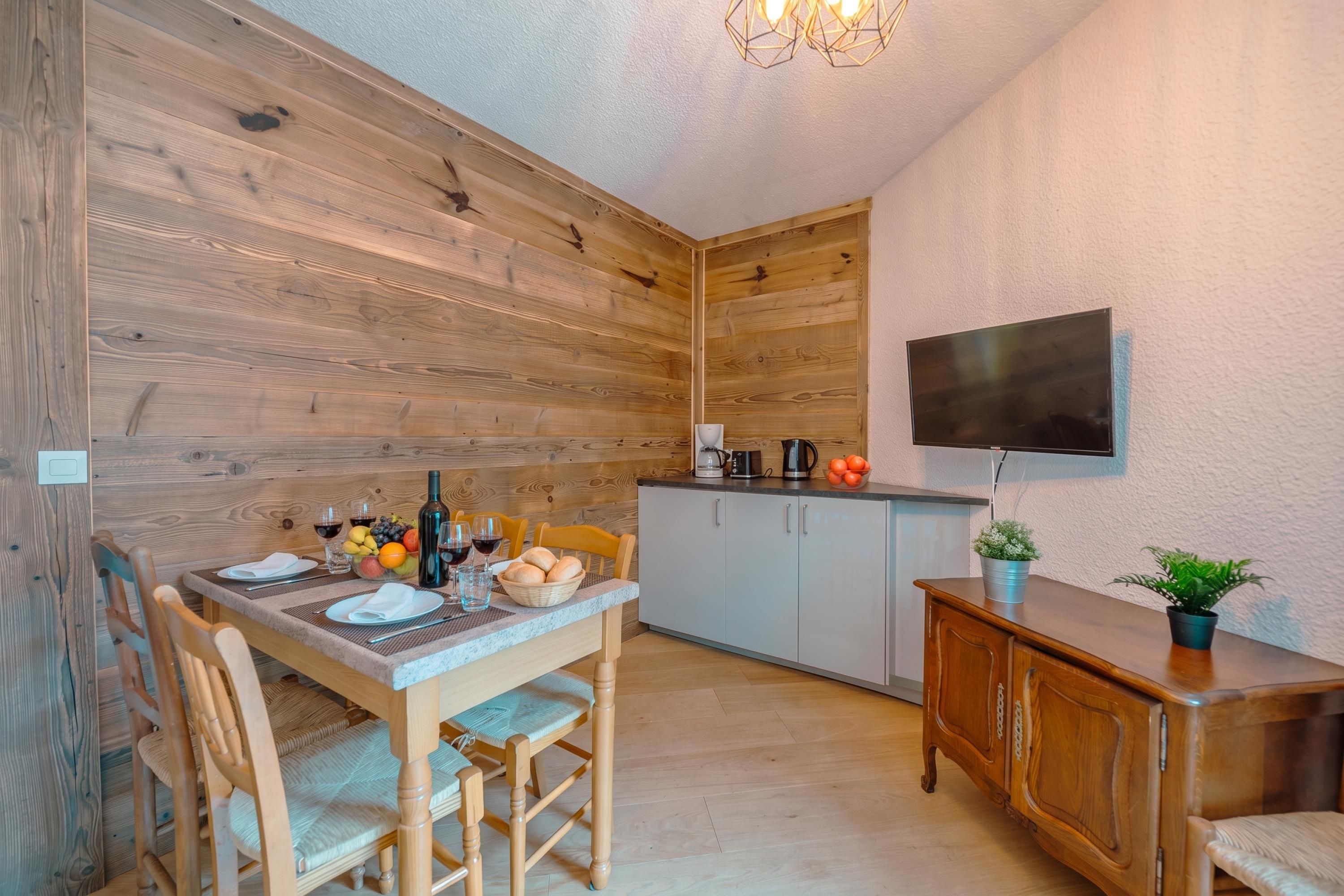 A wooden interior with a dining table set for two, a kitchen area with a TV, and a small cabinet with a plant.