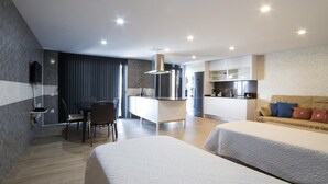 A modern hotel room with two beds, a dining area, a kitchenette, and a TV mounted on the wall.