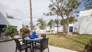 An outdoor patio with a round table and four chairs, surrounded by a grassy area with palm trees and a white building in the background.