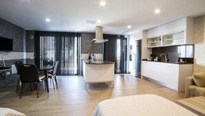 A modern hotel room with a large bed, a small kitchen area, a dining table, and a seating area.