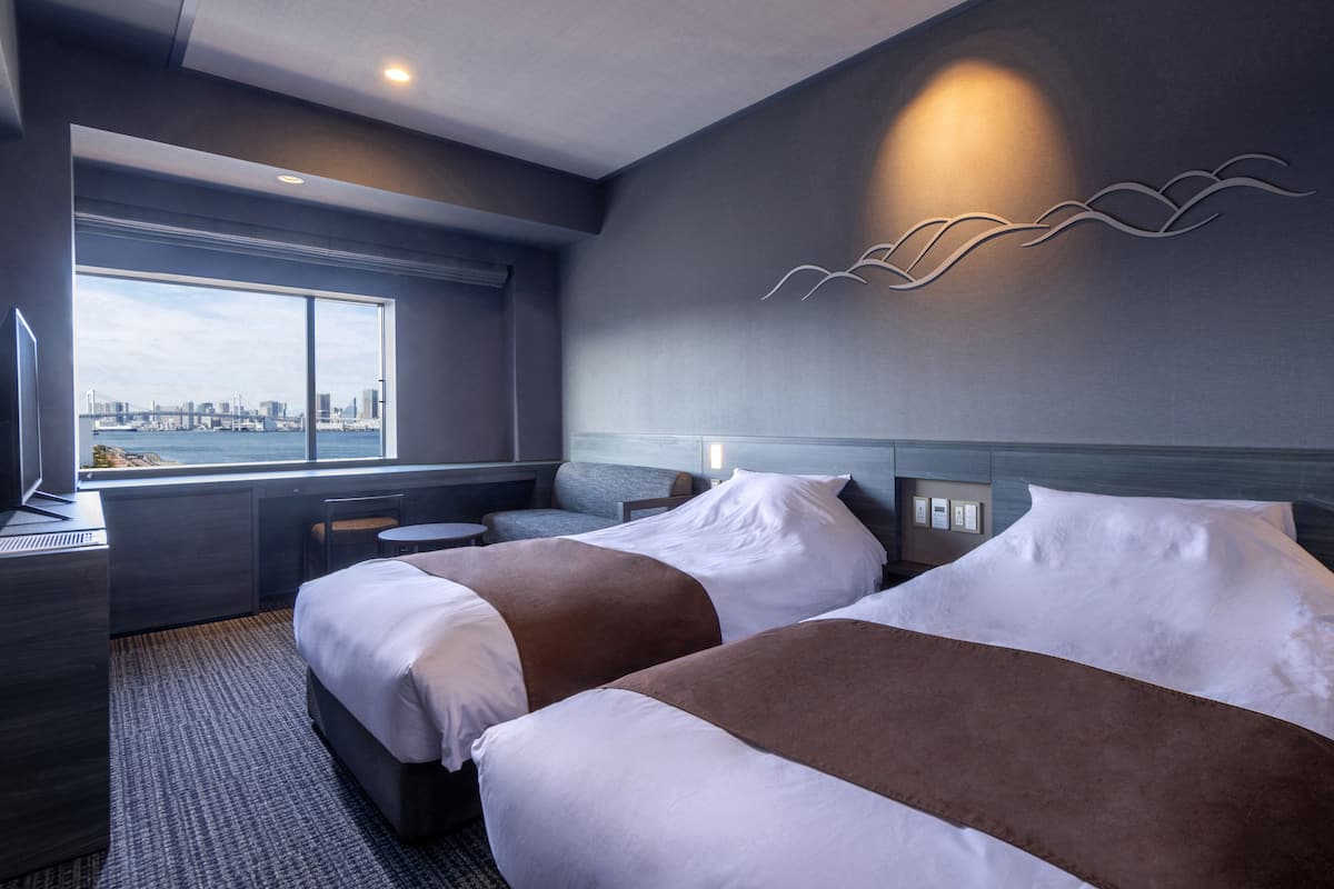 Habitación de hotel con dos camas, un escritorio y un amplio ventanal con vistas a la ciudad.