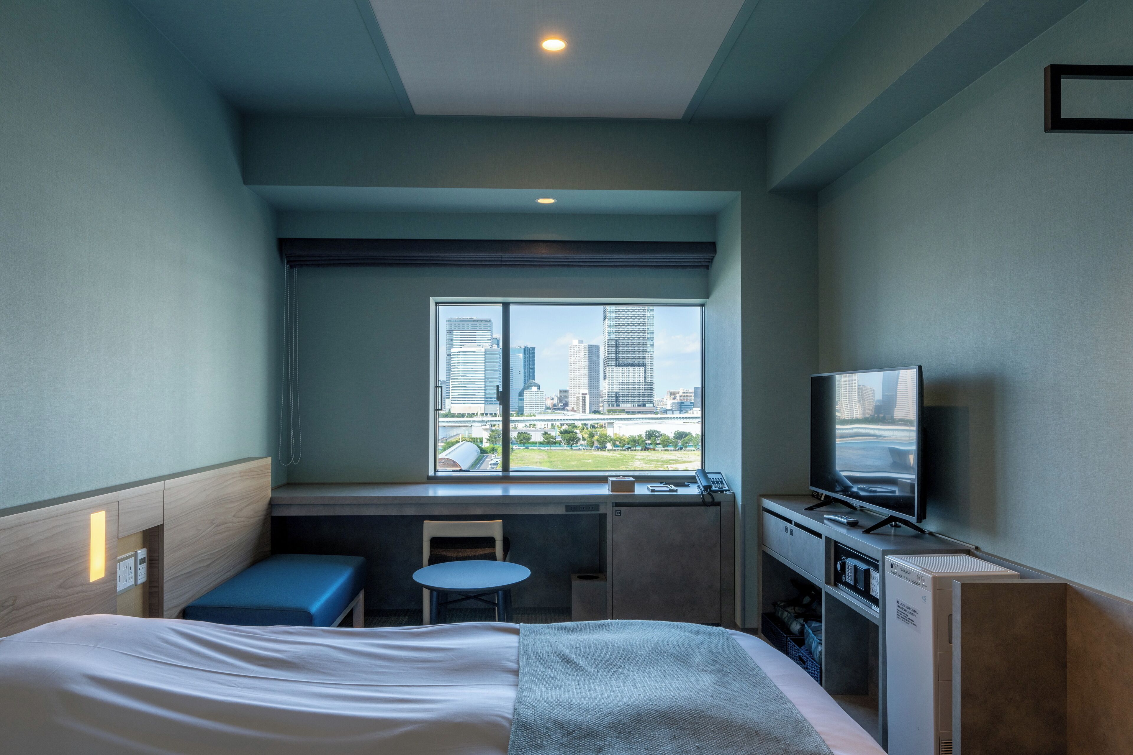 A hotel room with a bed, a desk, a chair, a TV, and a window with a city view.