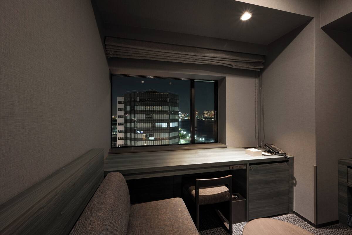 Habitación de hotel con escritorio, silla, banco y una amplia ventana que ofrece vista nocturna de la ciudad.