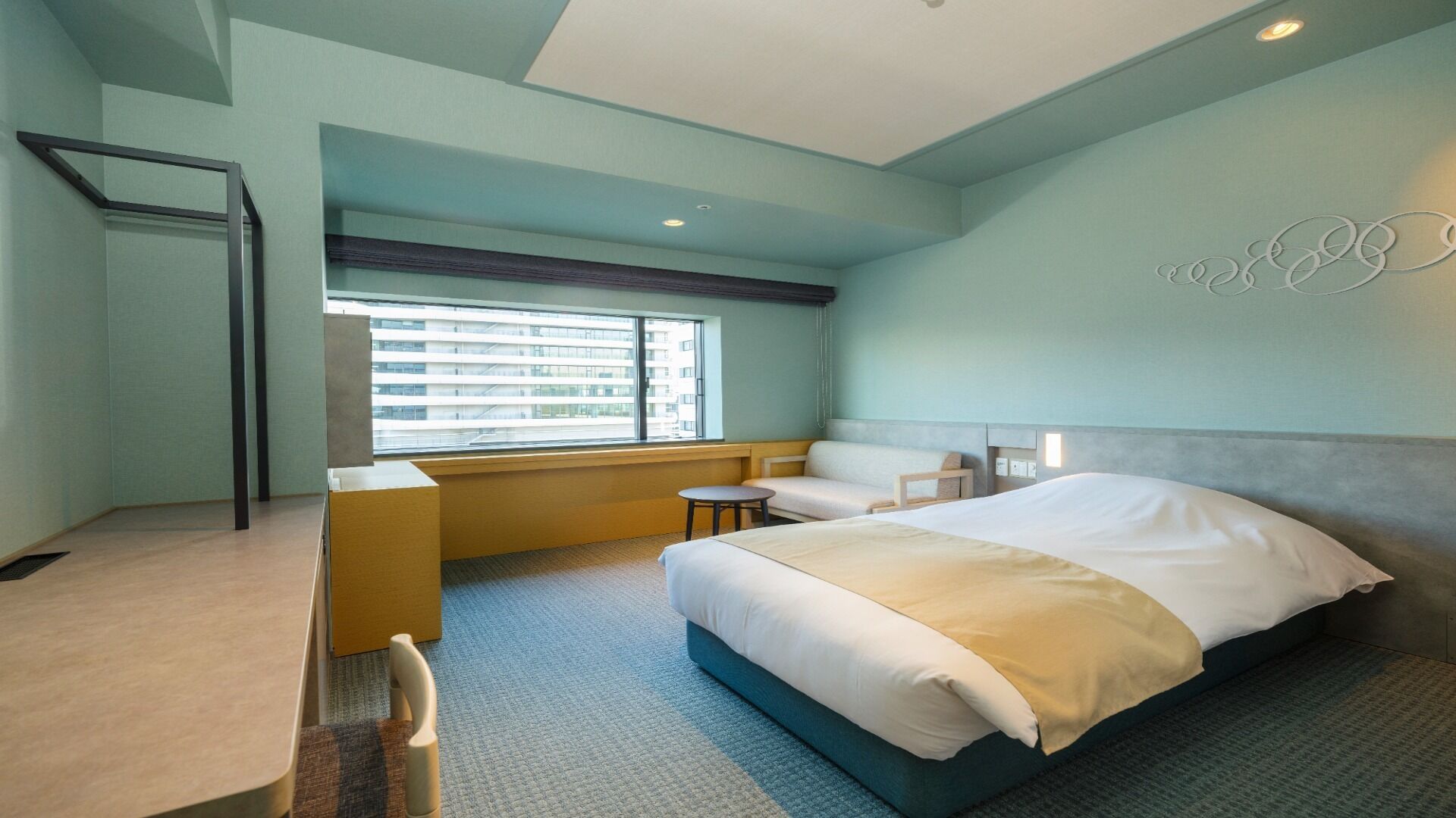 A hotel room with a bed, a desk, a chair, a window with blinds, and a wall with a circular design.