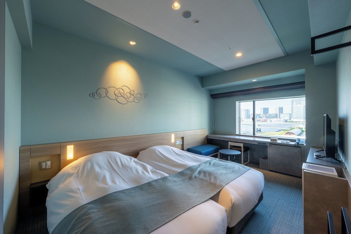 Habitación de hotel con dos camas, un escritorio, una silla, un televisor y una ventana con vista a la ciudad.