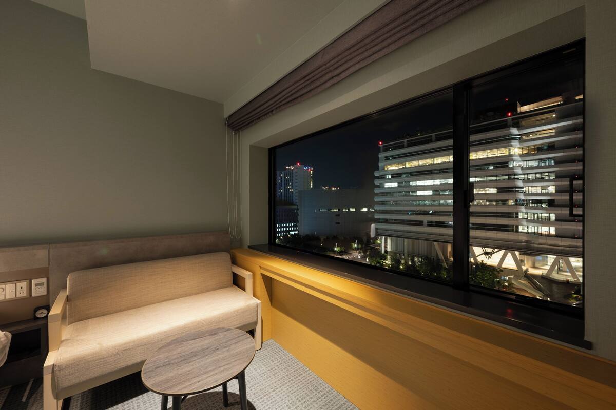 Una habitación de hotel moderna con sofá y una pequeña mesa, una ventana grande con vistas a un paisaje urbano y un puente por la noche.