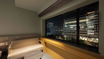 Una habitación de hotel moderna con sofá y una pequeña mesa, una ventana grande con vistas a un paisaje urbano y un puente por la noche.